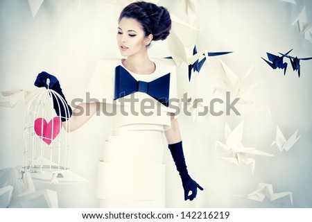 Art fashion photo of a gorgeous woman in paper dress holding birdcage with red heart. Black and white.