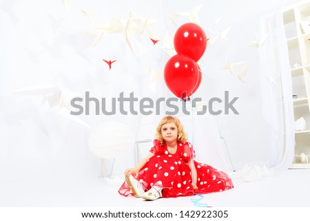 Cute little girl sitting with red balloons in a white room surrounded with paper birds.