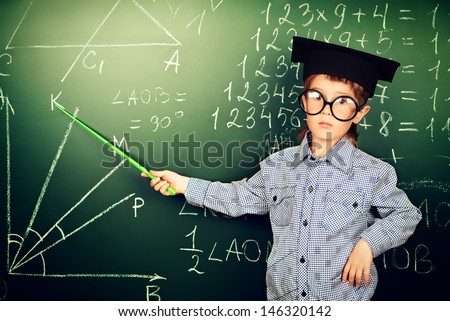Portrait of a boy in round glasses and academic hat standing near the blackboard in a classroom.