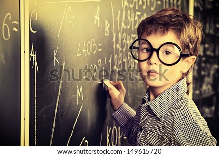 Portrait of a cute schoolboy in round glasses writing on a blackboard in a classroom.