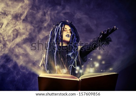 Little girl in a costume of witch casts a spell over magic book over dark background.