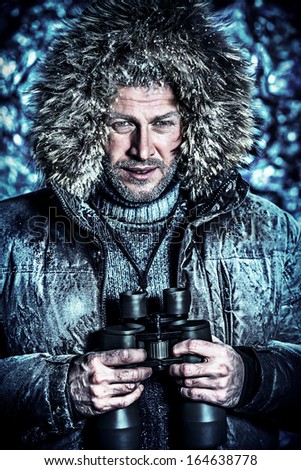 Handsome brutal man dressed in winter clothes covered with frost looking through binoculars.