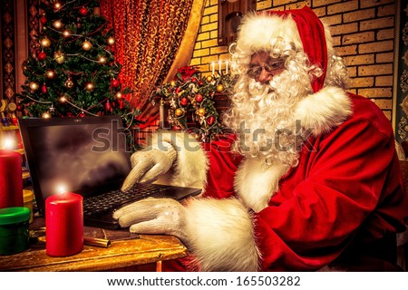 Santa Claus sitting at the table with a laptop at his home. Christmas time.