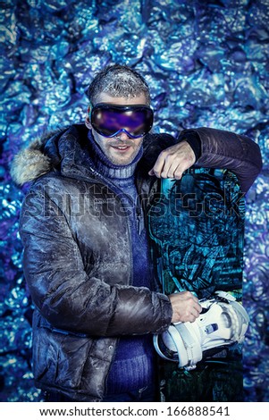 Smiling handsome man dressed in winter clothes, holding snowboard, covered with snow.