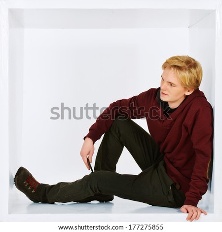 Young man in casual clothes sitting in white cube. Concept of rules and restrictions. Isolated over white.