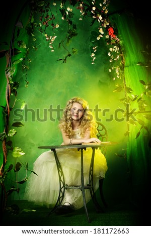Lovely girl in a lush white dress sitting at the table under a floral arch over green background.