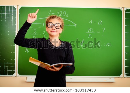 A schoolgirl in spectacles stands at the blackboard with an open book and holds up index finger. Education.