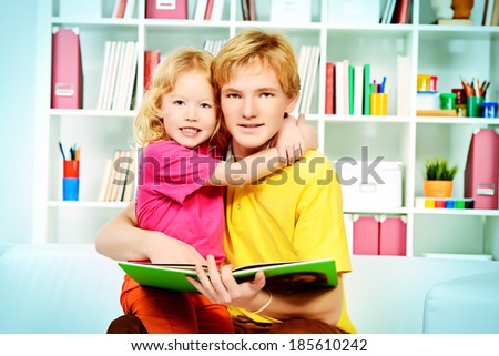 Elder brother and his little sister sitting together on sofa at home and reading a book.