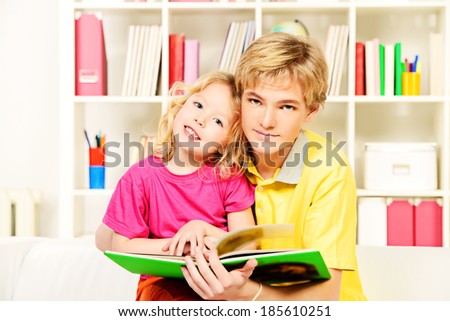 Elder brother and his little sister sitting together on sofa at home and reading a book.