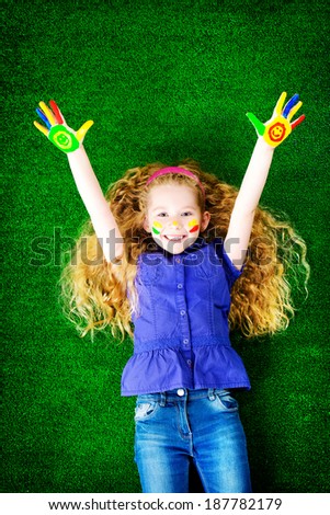 Laughing little girl painted in bright colors lying on green grass. Happy childhood.