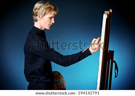Young man artist paints on canvas with oil paints. Studio shot over black background.