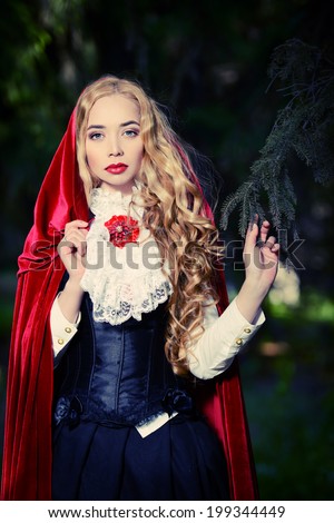 Portrait of a stunning blonde lady in  old-fashioned dress and red cloak in a fairy forest.