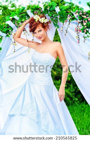 Beautiful bride with chaming red hair stands under the wedding arch. Wedding dress and accessories. Wedding decoration.