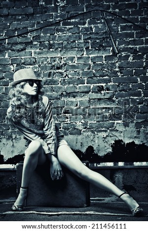 Portrait of a beautiful young woman sitting on her old suitcase near the brickwall. Fashion shot. Black-and-white photo.