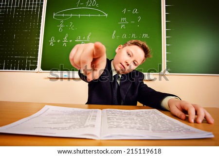 Smart schoolboy at a classroom points his finger at the camera. Education.