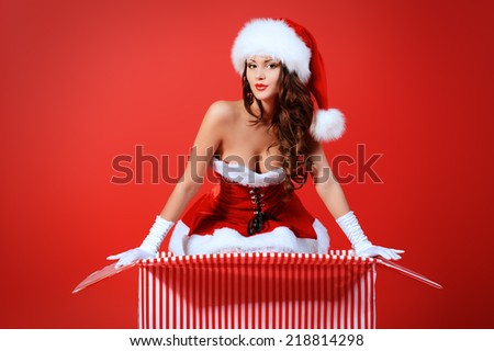 Beautiful young woman in Santa Claus costume posing with a huge gift box over red background. Christmas.