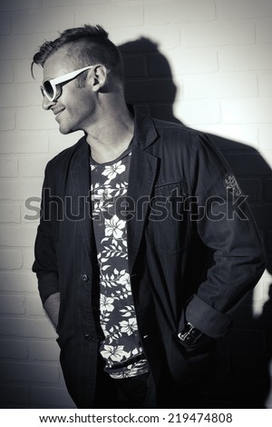 Black-and-white portrait of a stylish handsome man in a suit and sunglasses posing by the white brick wall. Men\'s beauty, fashion.