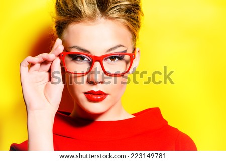 Close-up portrait of a stunning female model in red dress and elegant spectacles posing over yellow background. Beauty, fashion, optics.