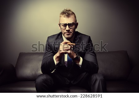 Portrait of a handsome mature man in elegant suit smoking a cigar. He is sitting on a leather couch in a luxurious interior.