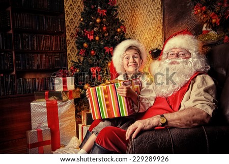 Happy little boy sitting on the lap of Santa Claus and rejoice a gift. Christmas decoration.