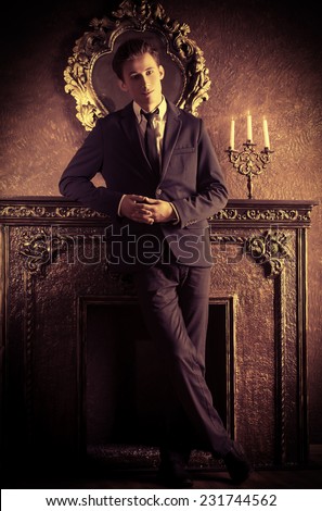 Young handsome man in elegant suit stands by the fireplace in a room with classic vintage style. Fashion.