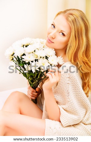 Pretty young woman sitting on a sofa at home, holding white flowers and smiling. Interior.