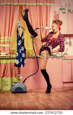 Charming pin-up girl ironing her dress and singing on a glamorous pink kitchen. Retro style. Fashion.