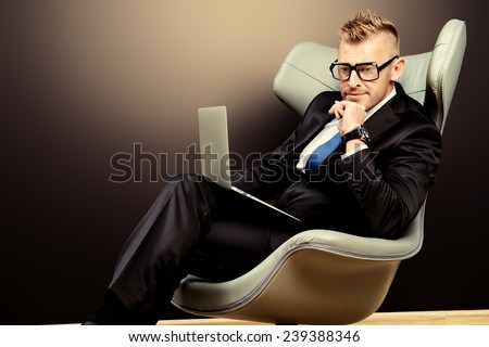 Imposing mature man in elegant suit sitting on a leather chair in a modern luxurious interior and working on a laptop. Fashion. Business.