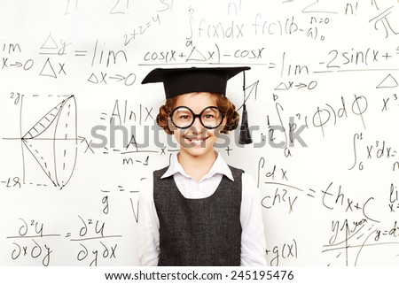 Happy smart schoolgirl in big glasses and academic hat performs the task at the blackboard. Education.