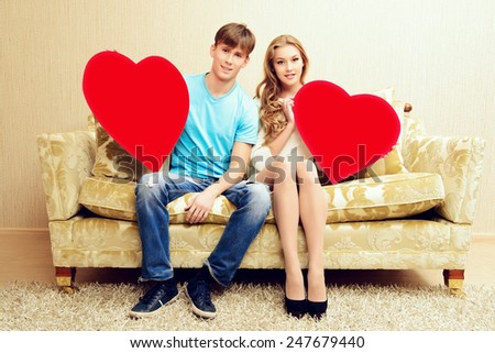 Romantic young couple sitting relaxed on a sofa in the cozy living room of their home. They hold big red hearts. Valentine day.