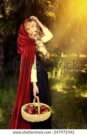 Beautiful blonde woman in  old-fashioned dress and red cloak walking throgh the forest with a basket of apples.