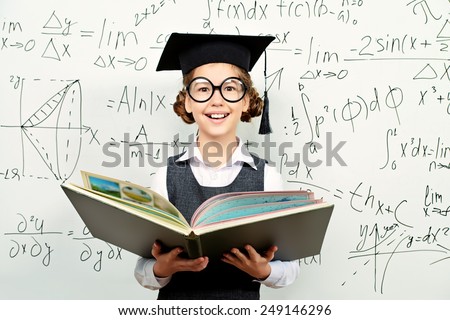 Pretty smart schoolgirl in big glasses and academic hat stands with opened book at the blackboard. Education.