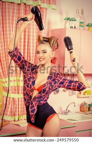 Beautiful pin-up girl doing a hair with a hair dryer and comb on her pink kitchen. Retro style. Fashion.