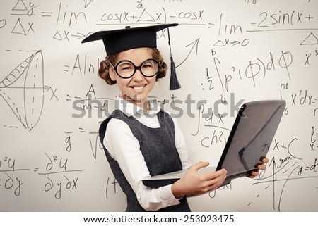 Smiling smart schoolgirl in big glasses and academic hat performs the task at the blackboard with a laptop. Education.