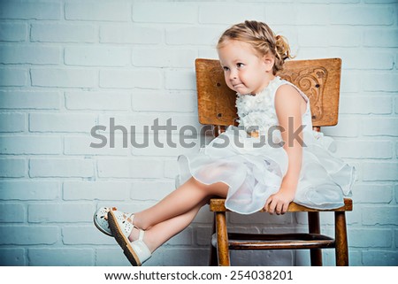 Sweet little girl in a beautiful white dress sitting on the old chair by the white brick wall. Childhood.