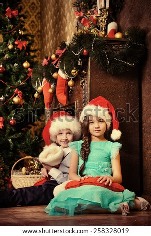 Two cute children in Santa hats sitting with gifts by the Christmas tree at home. The magic of Christmas.