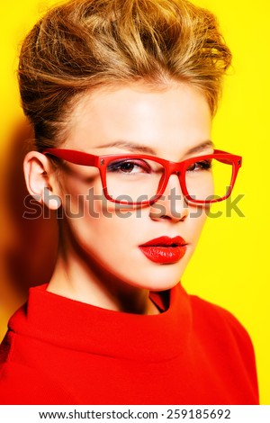 Close-up portrait of a stunning female model in red dress and elegant spectacles posing over yellow background. Beauty, fashion, optics.