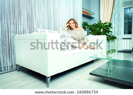 Smiling elegant woman sitting on a sofa in a living room. Home interior, furniture. Lifestyle.