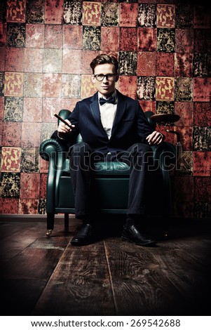 Handsome young man in elegant suit smoking a cigar. He is sitting on a leather chair in a luxurious interior.