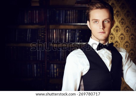 Young handsome man in evening suit stands by the fireplace in a room with classic vintage interior. Fashion.