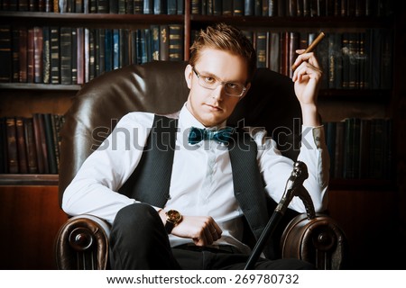 Elegant young man in a suit sitting in armchair and smoking a cigar. Vintage room. Fashion.