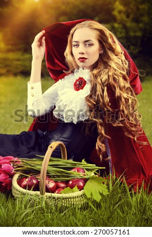 Beautiful blonde woman in  old-fashioned dress and red cloak sitting on the meadow with a basket of tulips.