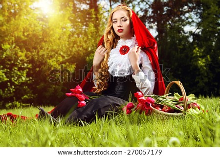Beautiful blonde woman in  old-fashioned dress and red cloak sitting on the meadow with a basket of tulips.