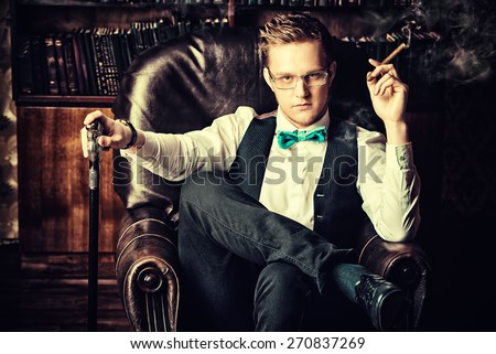 Elegant young man in a suit sitting in armchair and smoking a cigar. Vintage room. Fashion.