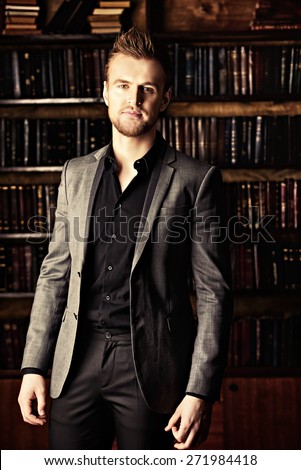 Handsome well-dressed man stands by bookshelves in a room with classic interior. Fashion.