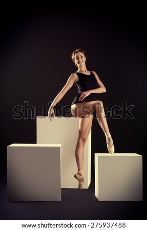 Elegant beautiful ballet dancer in black ballet leotard posing at studio on white cubes. Art concept.