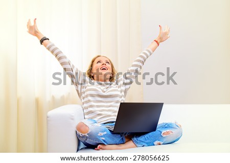 Young woman sitting on a sofa with her laptop computer raised her hands in the air, expressing happiness and success.