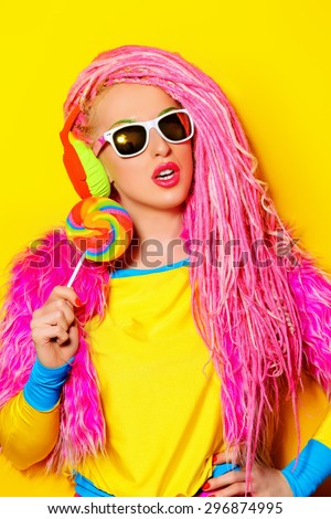 Attractive glamorous girl wearing ultra bright clothes and with pink dreadlocks eating lollipop. Bright style. Party style.