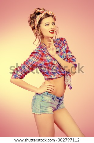 Attractive smiling pin-up girl alluring in shorts and shirt over pink background. Beauty, fashion.