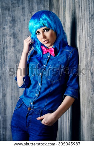 Trendy teen girl in bright blue wig and jeans clothes posing by a grunge wall. Beauty, fashion.